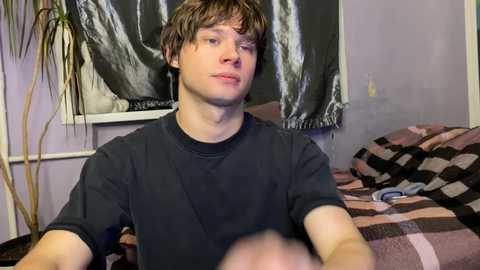Video of a young Caucasian male with messy brown hair, wearing a black t-shirt, sitting on a bed with a checkered blanket, in a dimly lit room with a black and silver curtain.