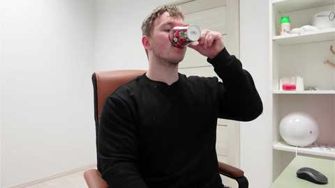 Video of a young, light-skinned man with short brown hair, drinking from a colorful can, seated in a tan leather chair in a white-walled, minimalist room with shelves and a white globe.