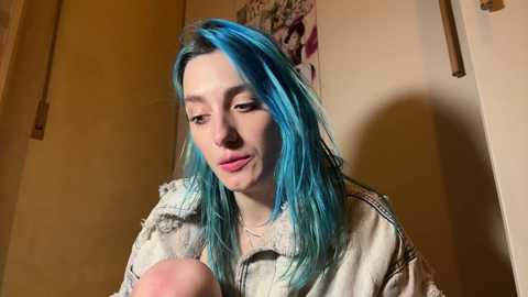 Video of a young woman with blue hair, wearing a beige jacket, looking contemplative while holding a small object. Background includes a cream-colored wall and a partially visible poster.