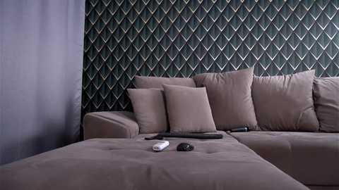 A video of a modern living room with a beige sectional sofa, two throw pillows, a remote control, and a television set. The wall features a dark green geometric patterned wallpaper.