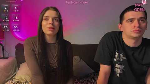 Video of a young couple on a bed, both wearing casual clothing; background shows a purple-lit room with a pillow and clock.