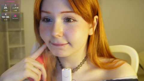 Video of a fair-skinned woman with long, straight, orange-red hair, wearing a black choker and off-shoulder top, smiling and touching her chin, in a cozy, warmly lit room.