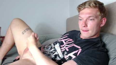 A video of a young, white, muscular man with short blonde hair and a tattoo on his thigh, lying on a bed in a T-shirt, legs spread, looking at a smartphone.