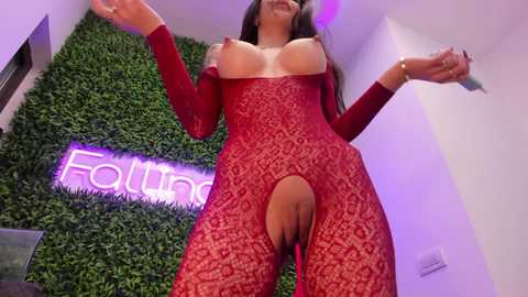 Video of a woman in a red lace bodysuit, exposing her large breasts and genitals, standing in a green foliage-themed room with a glowing \"Found\" sign.