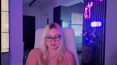 Video of a young, blonde woman with glasses, wearing a sleeveless top, seated in a modern kitchen with dim lighting.