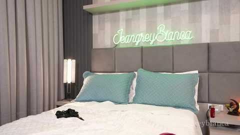 Video of a modern bedroom with a grey upholstered headboard, white bedding, turquoise pillows, and a neon sign reading \"DampneyBanana\" above.