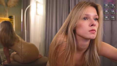 A video of a nude woman with long blonde hair, sitting on a bed in a dimly lit room. She has fair skin and is looking to the side.