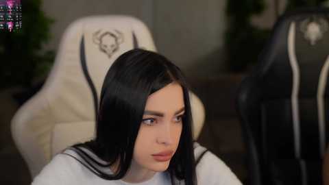 Video of a woman with long, straight black hair, wearing a white shirt, seated in a white gaming chair with a skull logo. Background shows a blurred, dark room with green plants.