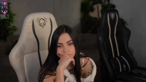 Video of a young woman with long black hair, wearing a white shirt and black jacket, sitting in a white gaming chair with skull design, holding her finger to her lips, in a dimly lit room.