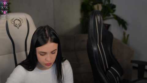 Video of a young woman with long black hair, wearing a white top, seated in a white gaming chair with a skull emblem. She's leaning forward, focused on a black gaming mouse. Background shows a beige sofa, potted plants, and a green wall.