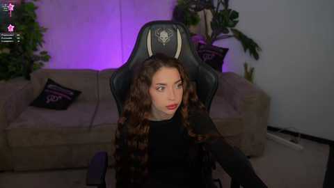 Video of a young woman with long, curly brown hair, wearing black, sitting in a gaming chair with a wolf logo, in a dimly lit living room with a beige sofa, potted plants, and purple lighting.