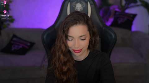 Video of a smiling woman with long, wavy brown hair in a black gaming chair, wearing a black top. Background features a grey couch, purple lighting, and a plant.
