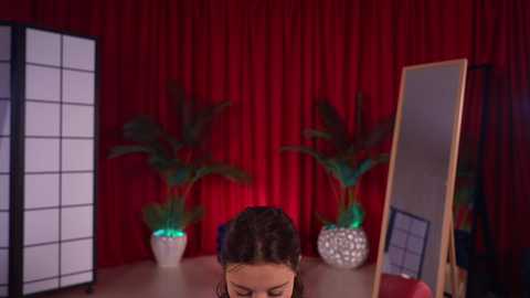 Video of a woman with dark hair, wearing a black top, sitting in a room with red curtains, green plants, and a mirror.