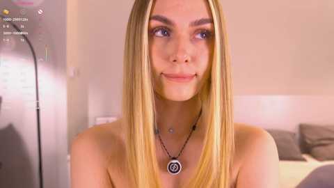 Video of a young, fair-skinned woman with long blonde hair, wearing a black necklace with a circular pendant. She smiles slightly, set against a blurred background with a bed and a lamp.
