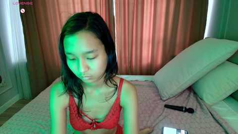 A video of a young Asian woman with long black hair, wearing a red lace bra, lying on a bed with beige sheets, green light illuminating her face.