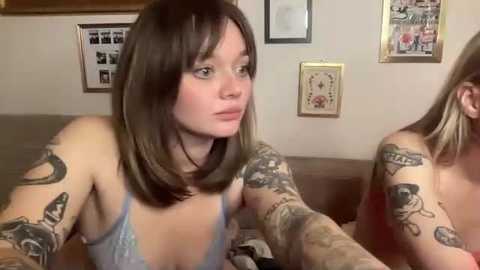 Video of two women with tattoos, one in a blue bra, the other in a red top, sitting on a couch in a room with framed art on beige walls.