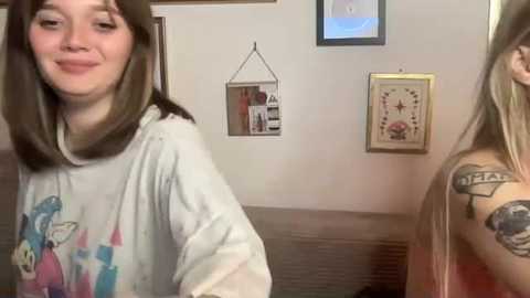 Video of two young women in a cozy room with white walls, a TV, framed artwork, and a gray couch. One woman has a floral shirt and a tattoo on her arm.