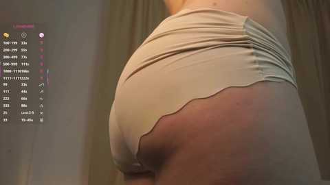 A close-up video of a person's buttocks clad in tight, beige shorts with a ruffled hem, standing against a blurred, neutral-colored background. A digital interface with a list of numbers and symbols is visible on the left.