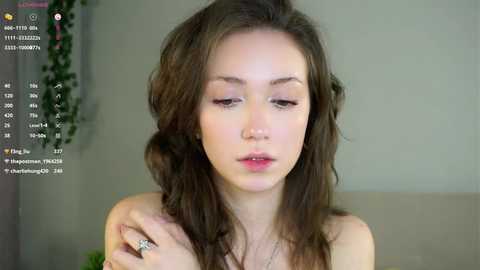 Media: Video of a young, light-skinned woman with wavy brown hair, wearing a simple necklace, sitting indoors with a blurred background.