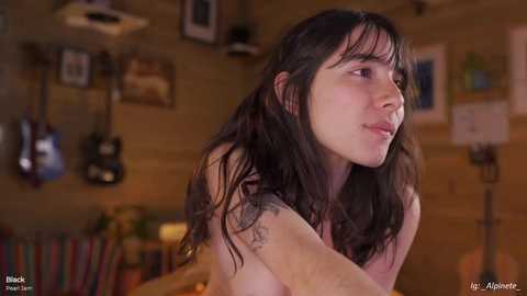 Video of a young, topless woman with long dark hair, sitting in a cozy, dimly lit room with wooden walls, guitars, and framed pictures.