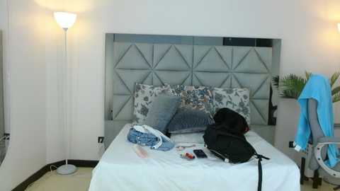 Video of a minimalist, modern bedroom with a grey geometric-patterned headboard, white walls, a white bed with blue pillows, a black backpack, a blue towel hanging on a chair, and a floor lamp.