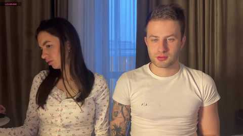 Video of a Caucasian man and woman in a room with beige curtains, the woman has dark hair, floral top, and is looking away, while the man with short hair and a white t-shirt looks serious.