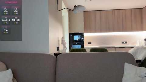 Video of a modern living room with a gray sofa, wooden kitchen cabinets, a digital clock, and a minimalist decor featuring a white statue and a green chair.
