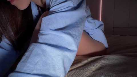Video of a woman in a blue shirt, lying on her stomach on a bed with a patterned beige blanket, her face partially visible, and the background showing a white wardrobe.