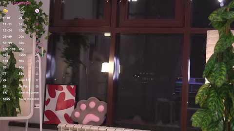 Video of a cozy, dimly-lit room with a large window, a red heart-patterned pillow, a paw print cushion, and green plants.