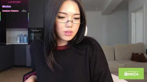 Video of a young East Asian woman with long black hair, wearing glasses, a black top, and a maroon collar. She's in a modern, minimalist living room.