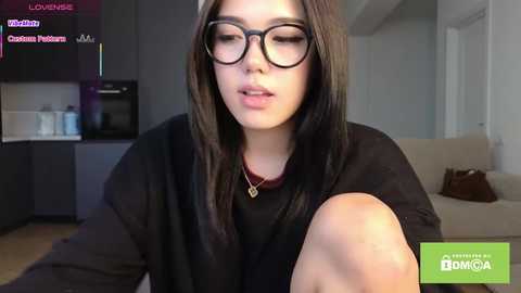 A video of a young woman with long brown hair, wearing glasses and a black sweater, seated in a modern living room.