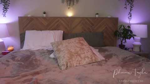 Video of a cozy, dimly-lit bedroom with a wooden chevron headboard, beige bedspread, and two white pillows. Two potted plants flank the bed, and hanging plants add greenery. Warm, purple-toned ambient lighting enhances the serene atmosphere.
