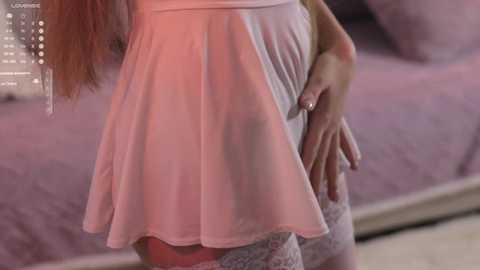 A video of a pregnant woman with fair skin and long, wavy brown hair, wearing a light pink, flowy dress and white lace stockings, standing in a dimly lit bedroom with a bed and a calendar in the background.