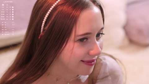 Video of a young Caucasian woman with long, straight brown hair, wearing a white pearl headband and a white dress, smiling softly. Background shows a blurred, light-colored room.