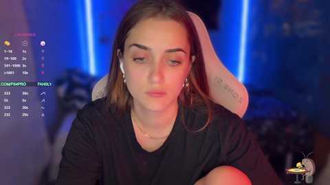 A video of a young woman with fair skin, brown hair, and a contemplative expression, wearing a black top, seated in a gaming chair with blue and red lights in the background.