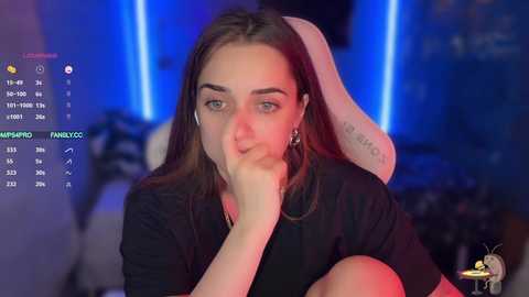 Video of a young woman with light skin and long brown hair, wearing headphones, sitting in a dimly lit room with blue and red lighting.