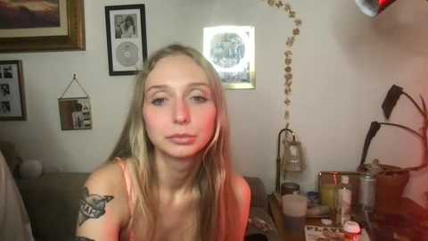 Video of a young blonde woman with a tattoo on her shoulder, wearing a pink tank top, in a dimly lit room with framed photos, a mirror, and a cluttered desk.