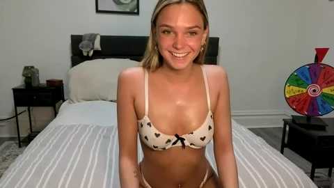 Video of a smiling, light-skinned, blonde woman in beige lingerie with heart patterns, sitting on a bed in a minimalistic bedroom with a colorful pinball machine.