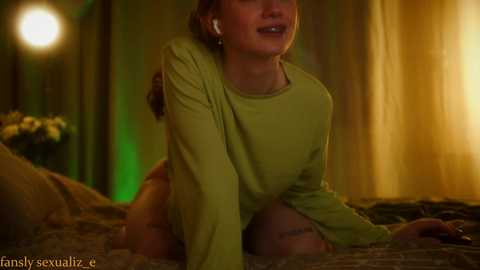 Video of a woman with light skin, long brown hair, and fair complexion, wearing a green long-sleeve shirt, kneeling on a bed with beige sheets, in a dimly lit room with green and yellow ambient lighting.