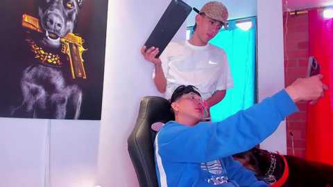 Video: Two young men, one in a blue jacket, the other in a white shirt, pose in a brightly lit room with a large, black and white dog portrait on the wall.