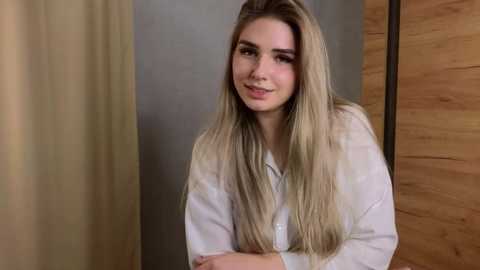 A video of a young Caucasian woman with long, straight blonde hair, wearing a white button-down shirt, standing in a modern, wooden-paneled room with beige curtains. She has a calm expression.