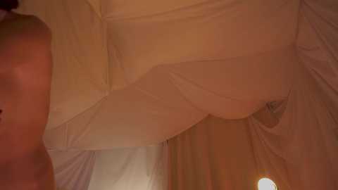 Video of a woman's legs in sheer white stockings, partially covered by a sheer white sheet, revealing a hint of her bare thigh. The background is a warm, dimly lit room with soft lighting.