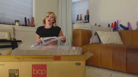 Video of a blonde woman in a black top, excitedly unwrapping a large brown box filled with sex toys in a modern living room.