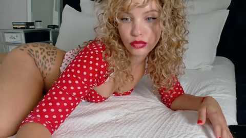 Video of a blonde woman with curly hair, wearing red polka-dot top and leopard-print panties, lying on a white bed with pillows, in a modern bedroom.