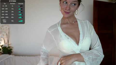 Media: Video of a smiling, fair-skinned woman in a sheer, white lace robe with a plunging neckline, standing in a bedroom with a white bed, orchid, and wooden door.