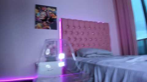 Video of a modern, minimalist bedroom with a pink, tufted headboard, a clear plastic chair, a neon purple strip, and a colorful poster on the wall.