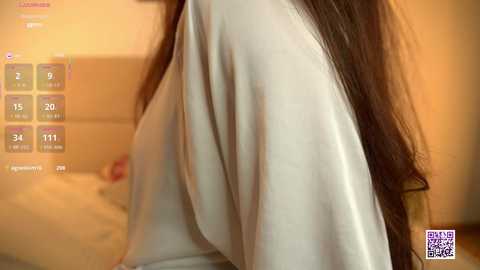 Video of a woman with long, dark hair, wearing a white robe, sitting on a bed. Background shows a warm, beige wall with a blurred clock displaying 2:15 AM.