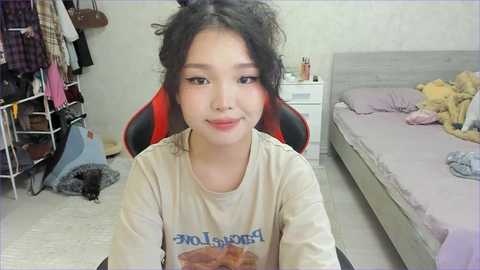 Video of an East Asian woman with curly black hair, wearing a cream t-shirt with a cartoon character, sitting on a red gaming chair in a messy bedroom with an unmade bed, clothes hanging, and a black cat.