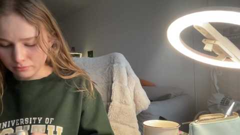 Media: Video of a young woman with light brown hair, wearing a green University of Michigan sweatshirt, sitting at a cluttered desk with a bright ring light, a blanket, and scattered papers in a dimly lit room.