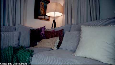 Video of a cozy living room with a grey sofa adorned with various patterned and textured pillows, a large white lamp, and a framed religious painting on the wall.
